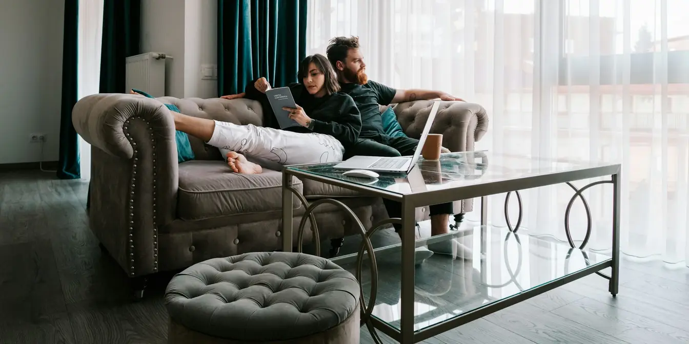 Two people relaxing on a sofa in a bright living room, one reading, a laptop on the coffee table