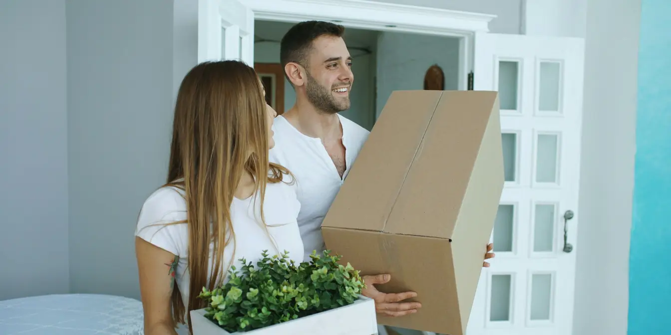 A couple carrying a cardboard box and a crate of plants through the door of a new home