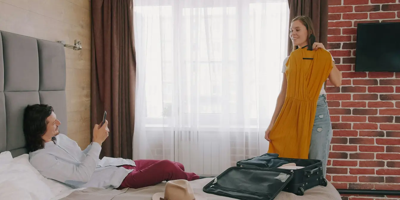 A woman holds up a yellow dress beside an open suitcase while her partner checks his phone on the bed