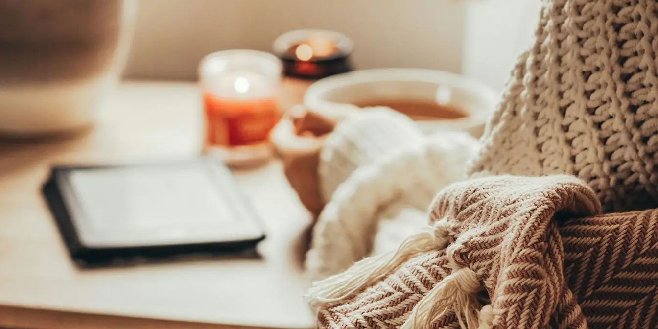 A cup of tea, lit candles and an e-reader on a table by a window, beside a knitted blanket