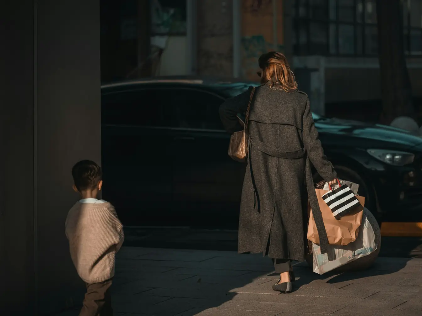 A woman in a long wool coat walks toward a parked car with an armful of shopping bags, a small boy following in the late-afternoon sun