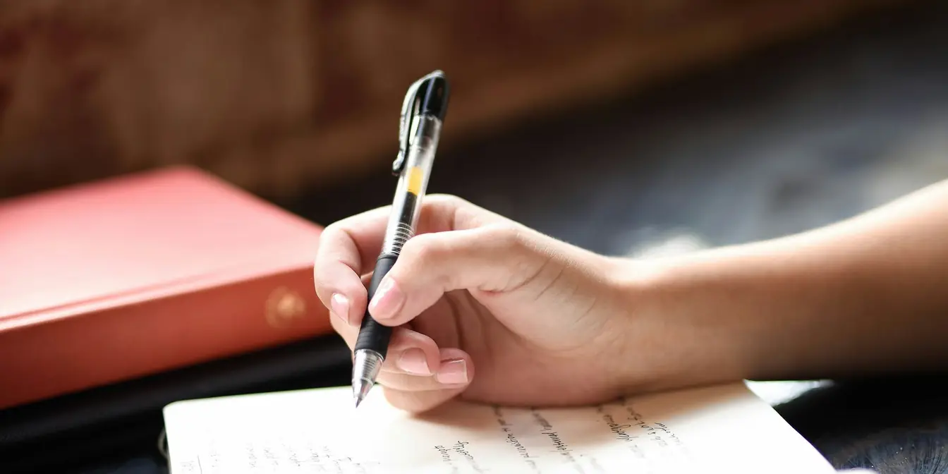 A hand writing a list in an open notebook at a table by a window